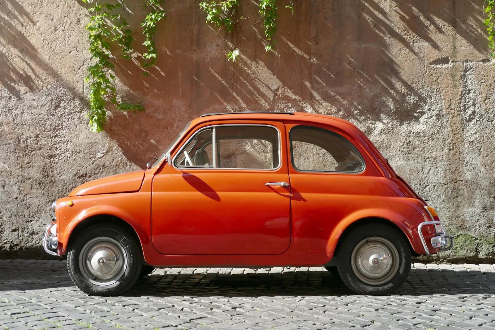 A vintage orange Fiat 500 parked on a cobblestone street against a weathered wall.